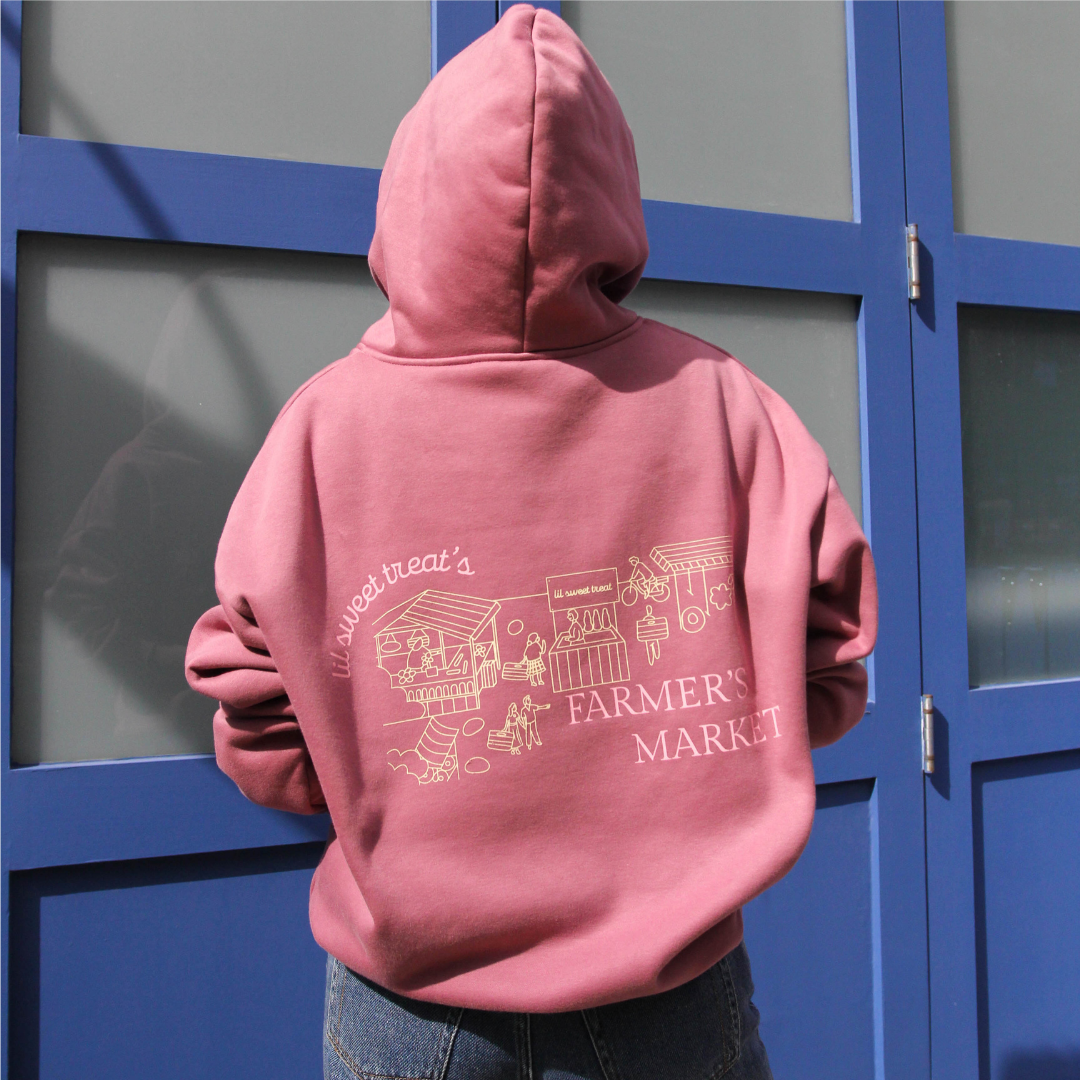 Person wearing a pink hoodie with 'Farmers Market' design in front of blue doors.